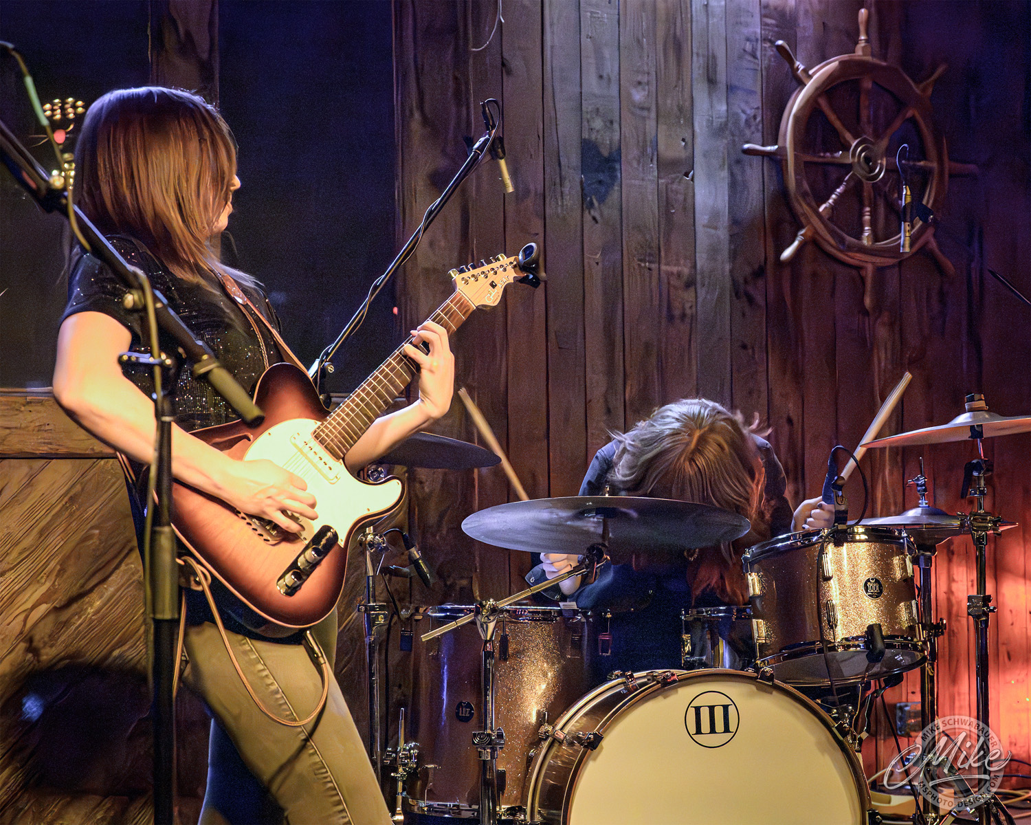 Two young women playing guitar and drums on a stage with wooden walls and nautical decor.