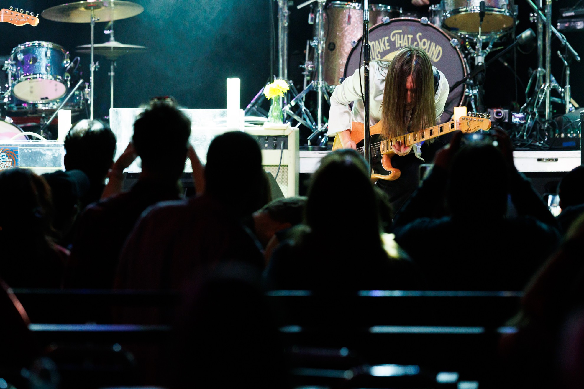 Musician with long hair playing guitar on stage with drums and equipment, audience watching, dim lighting, concert setting.