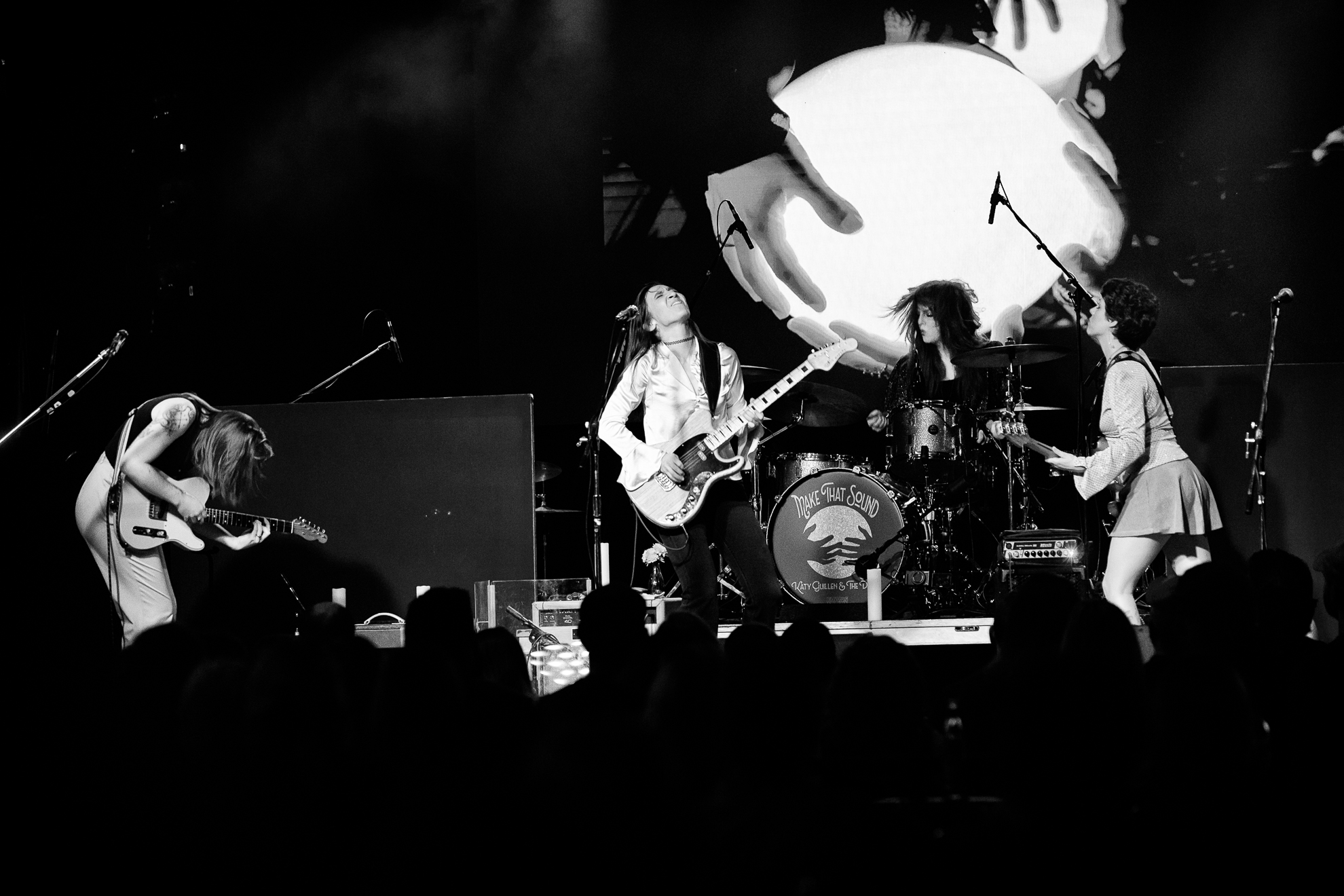 Band performs on stage with three musicians playing guitars and drums, audience visible in foreground, black and white image.