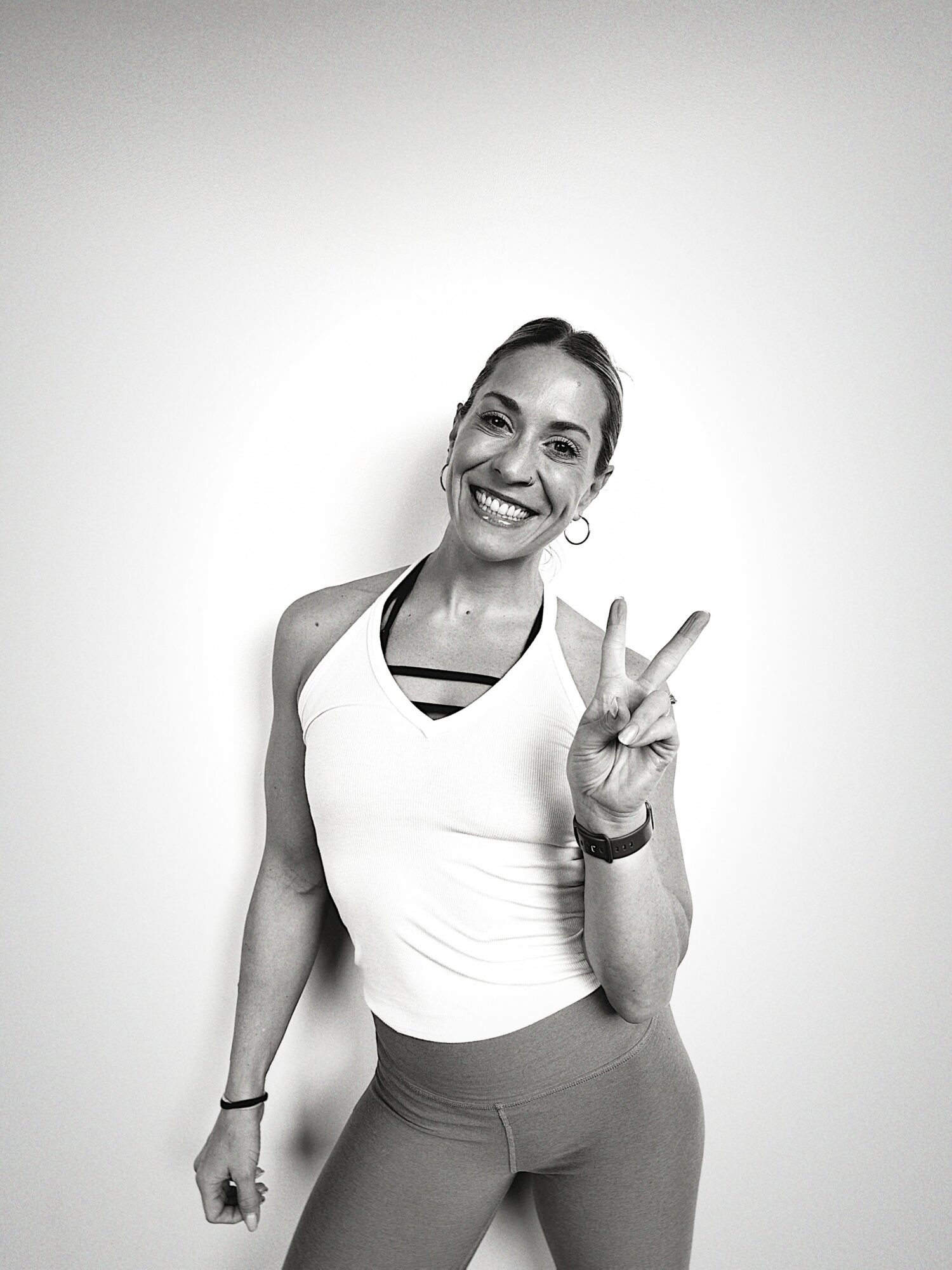 Smiling woman making a peace sign with her right hand, wearing a tank top and leggings, standing against a plain background.