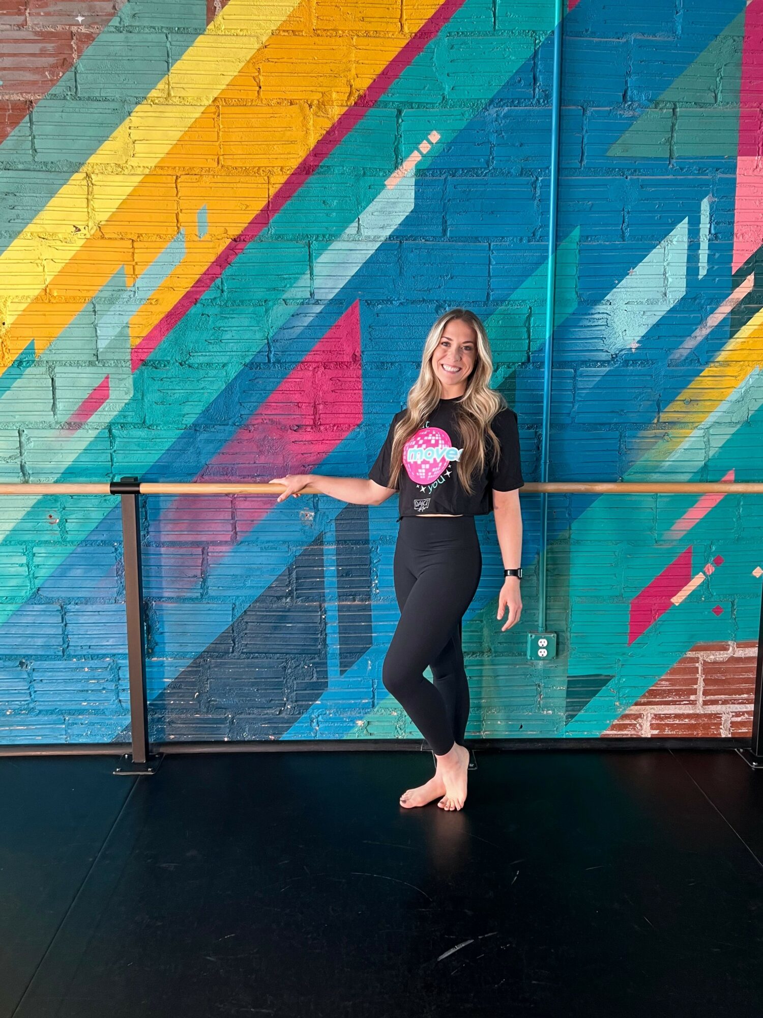 Person with long hair standing sideways against a colorful graffiti wall, holding a pink object, barefoot, with one foot on the ground.