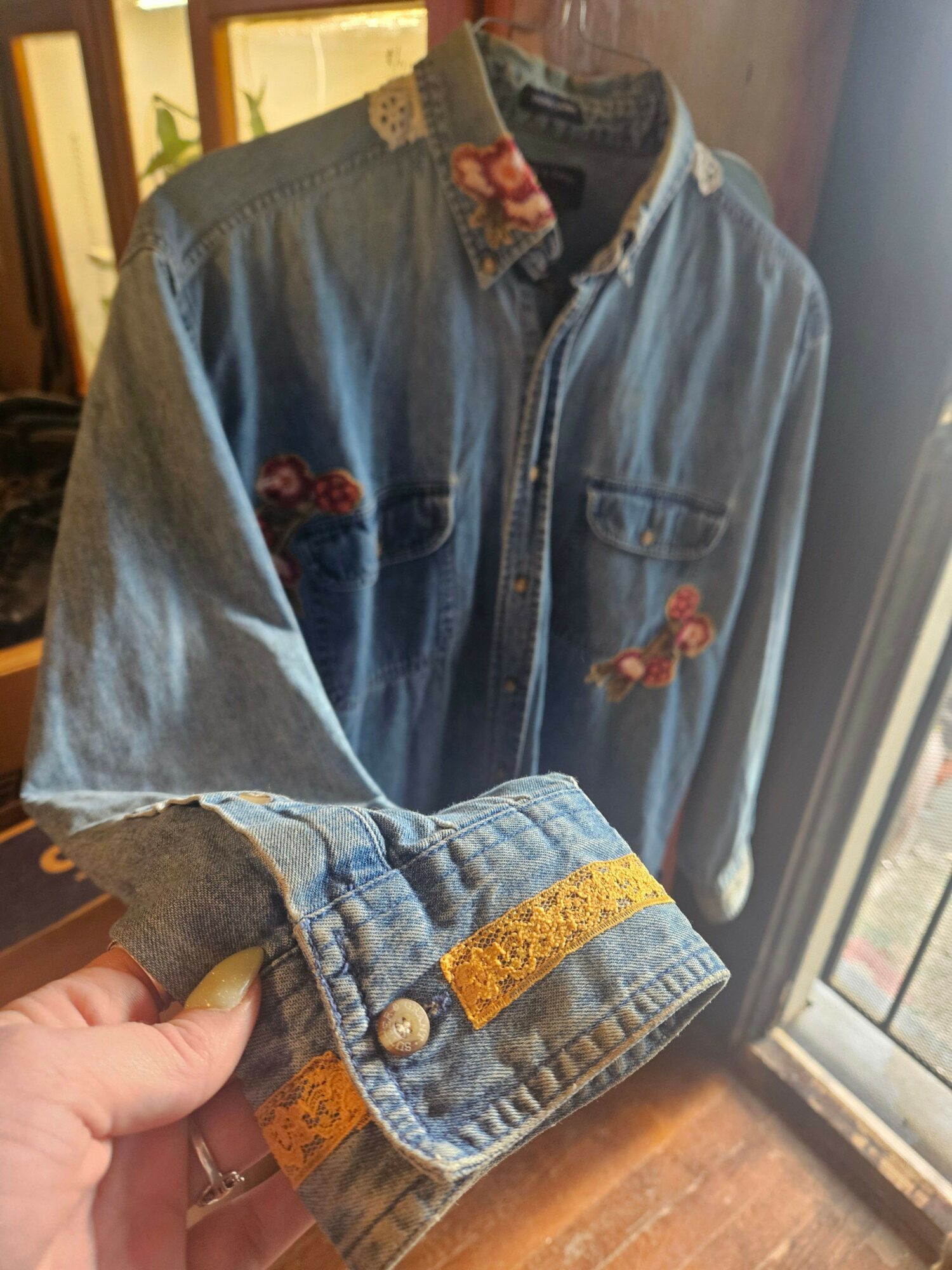 Close-up of a denim jacket with embroidered flowers, held by a person's hand near a window, sunlight illuminating the fabric.