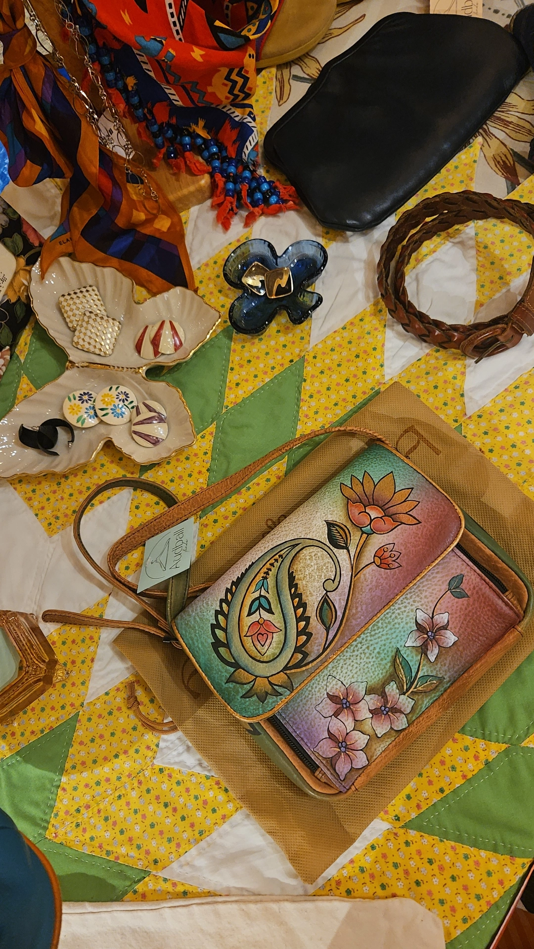 Colorful fabric items, a black case, a woven bracelet, and a floral purse on a yellow and green quilted surface.