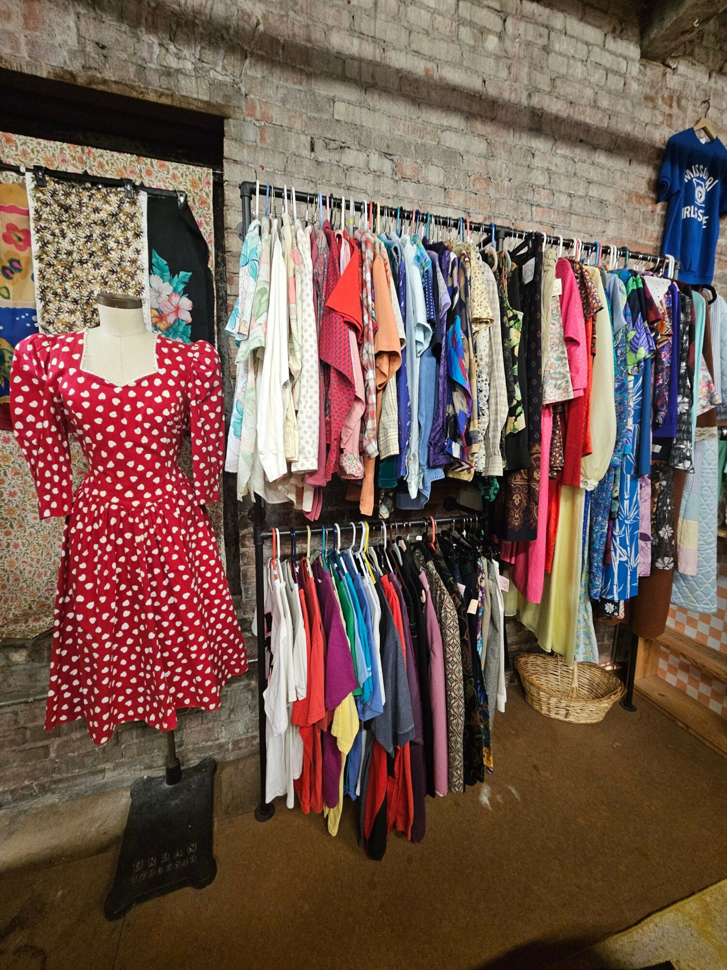 Stack of colorful folded clothes on a table, with a red polka dot dress hanging in front, and a black object nearby.