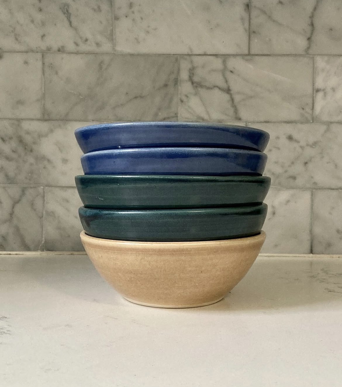 Stacked ceramic bowls in blue, green, and beige on a white surface with a marble wall background.