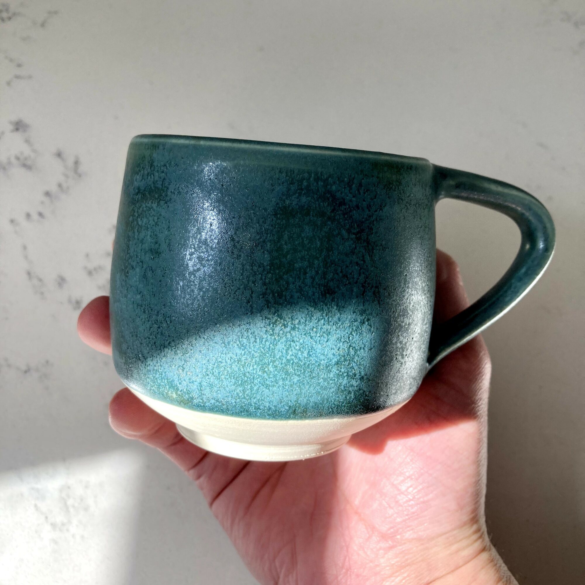 Hand holding a ceramic mug with a gradient blue-green glaze, against a light background.