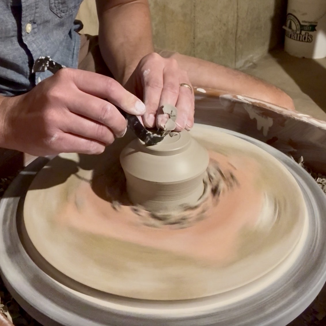 Person shaping clay on a pottery wheel with hands close-up, spinning clay on wheel, background with a bucket and wall.