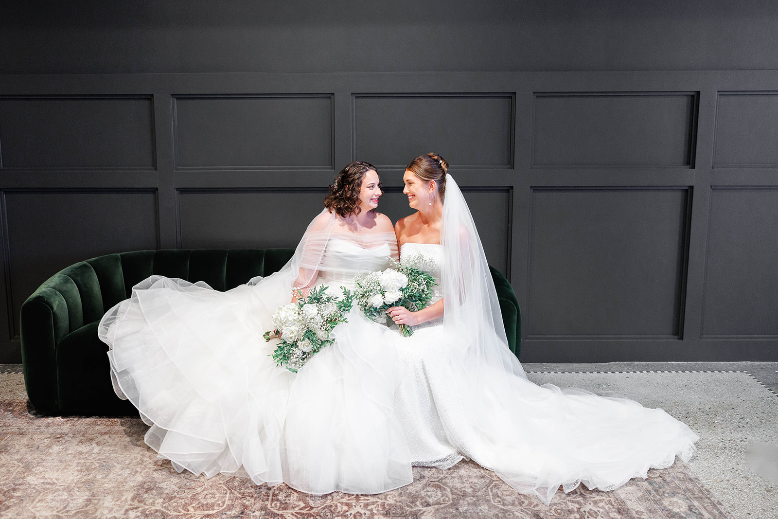 Two women in wedding dresses sit on a dark green sofa, holding bouquets, in front of a dark wall.