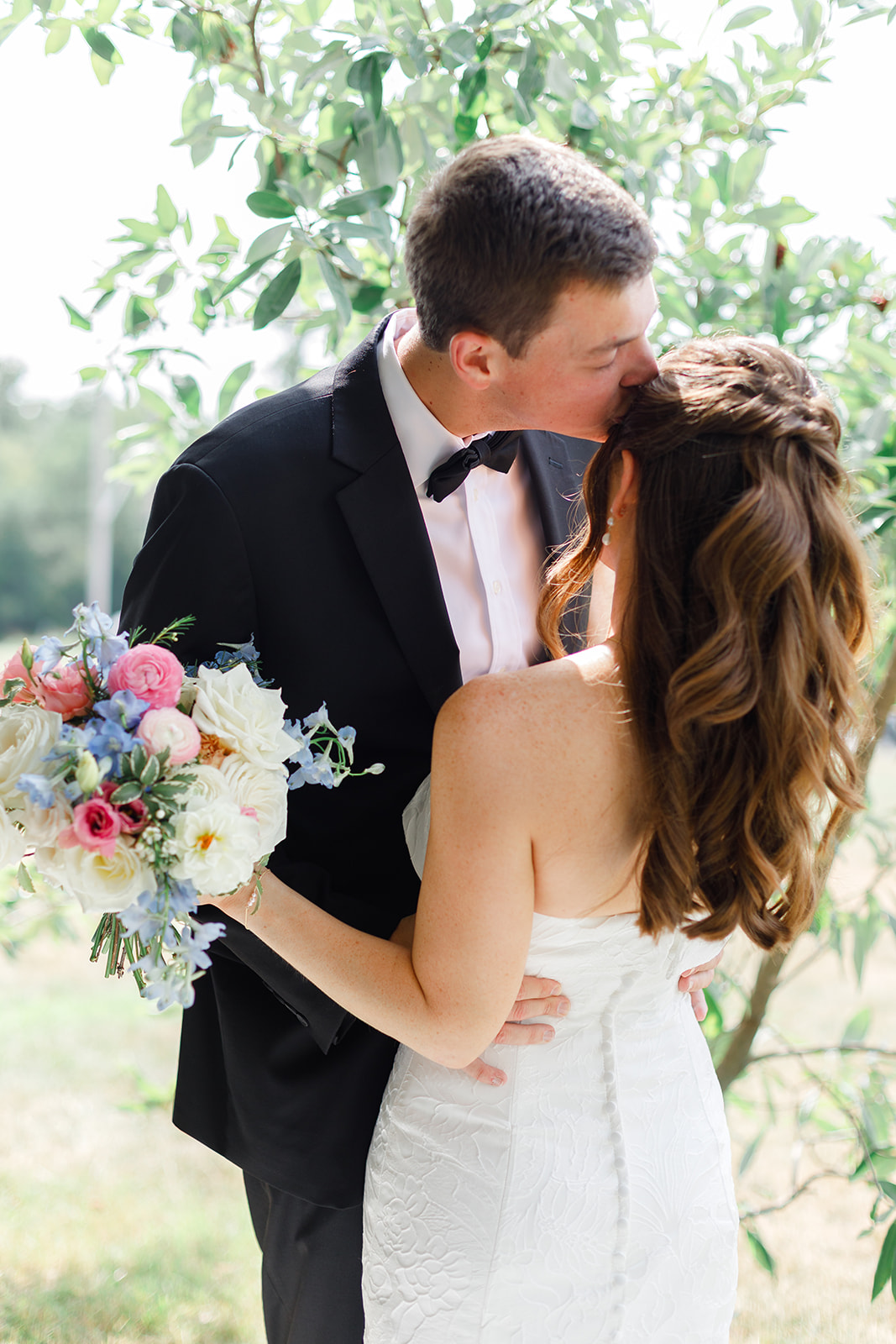A couple in wedding attire sharing a kiss outdoors, with a bouquet of flowers visible, surrounded by greenery.