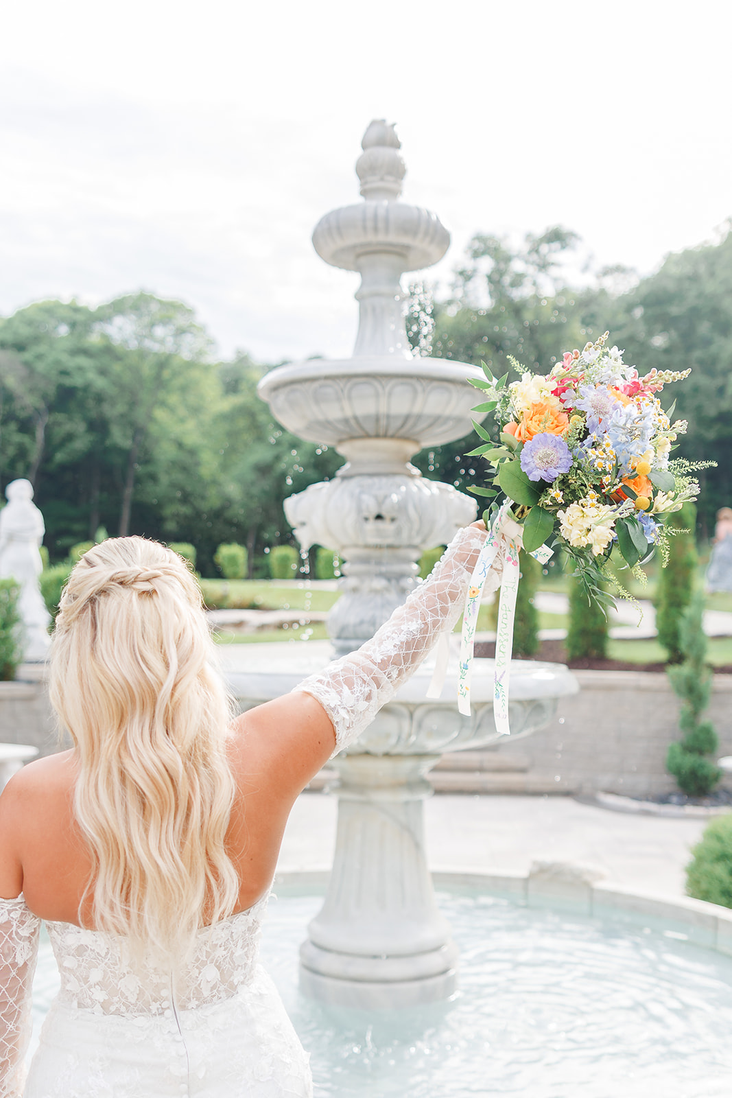 Woman in wedding dress holding a tray with flowers near a fountain in a garden.