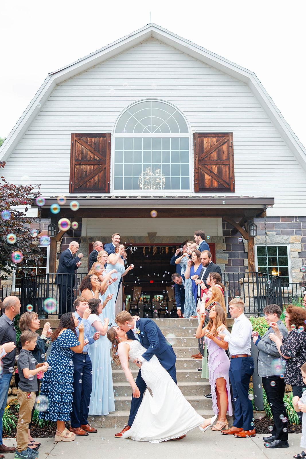 Wedding celebration outside a barn with guests cheering and blowing bubbles, bride and groom in the center, people in formal attire.