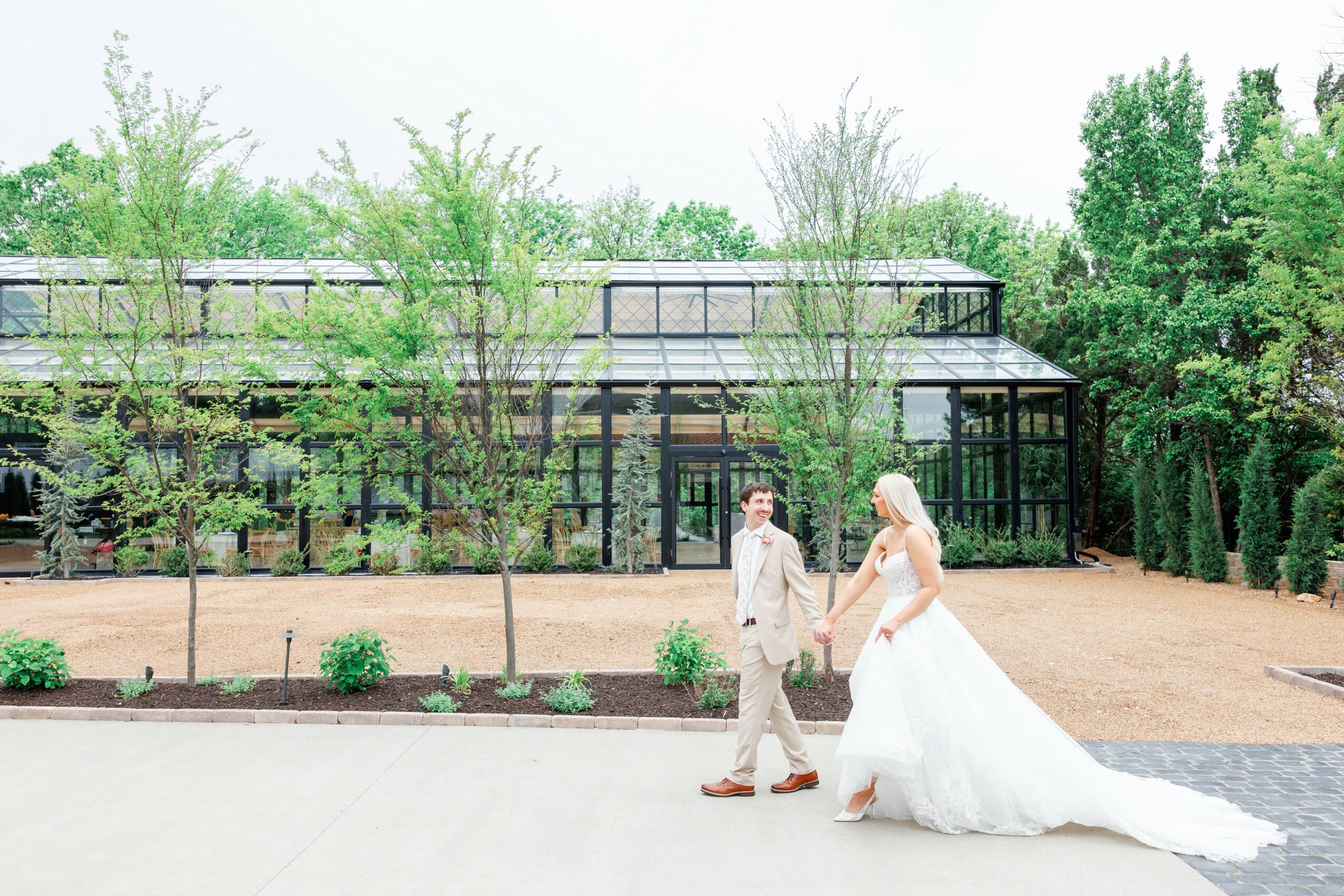 A bride and groom walk outdoors near a glass greenhouse, surrounded by trees and greenery, on a wedding day.