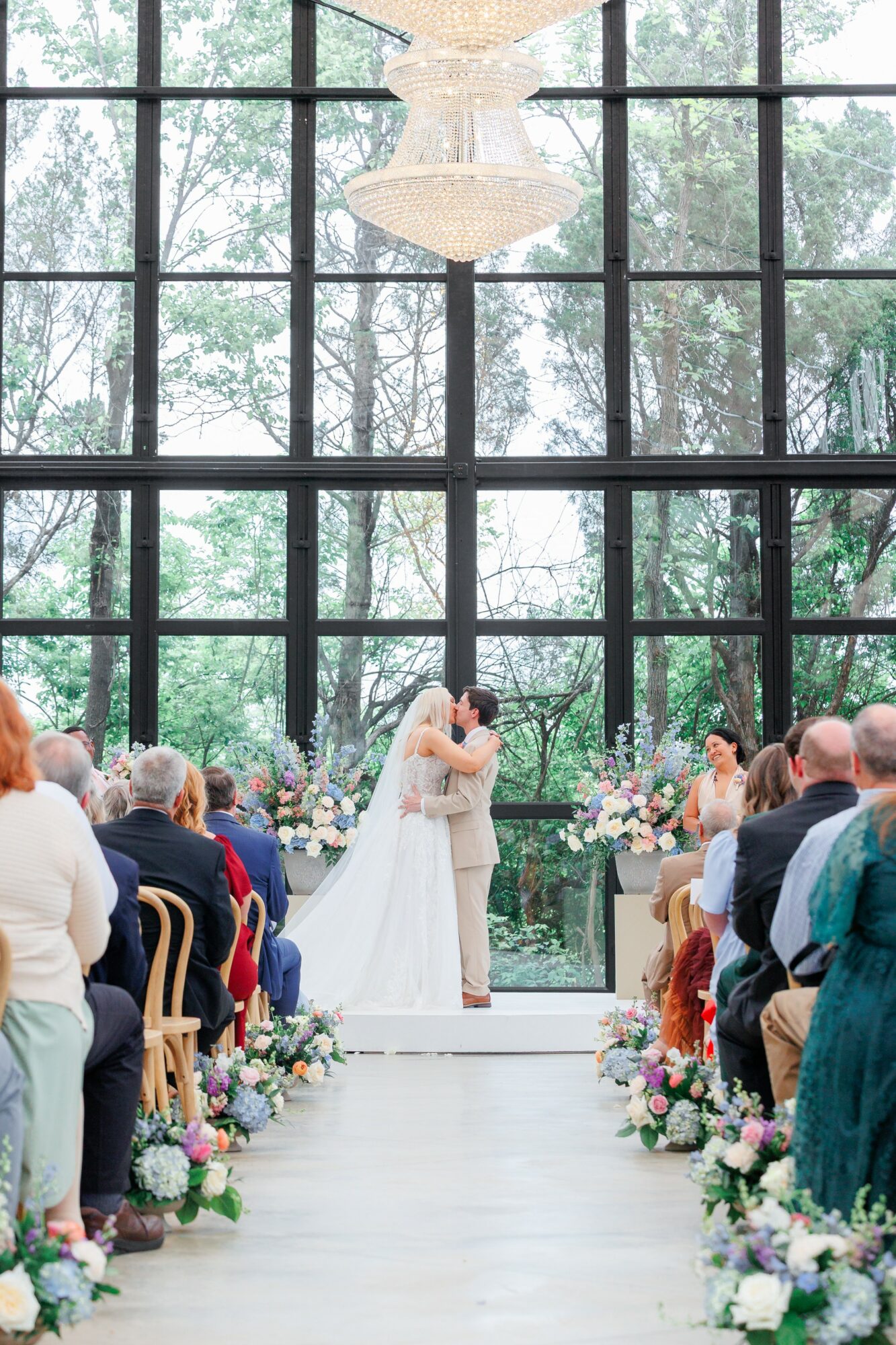 Couple kissing at wedding altar with guests seated on both sides, floral arrangements, large window, and chandelier above.