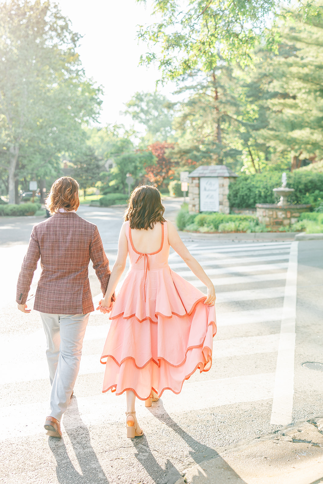 Two people walk across a crosswalk in a park with trees and greenery in the background.