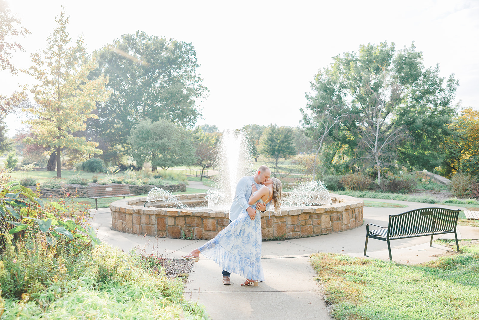A couple dancing in a park near a fountain with trees and benches, during daytime, with sunlight filtering through the trees.
