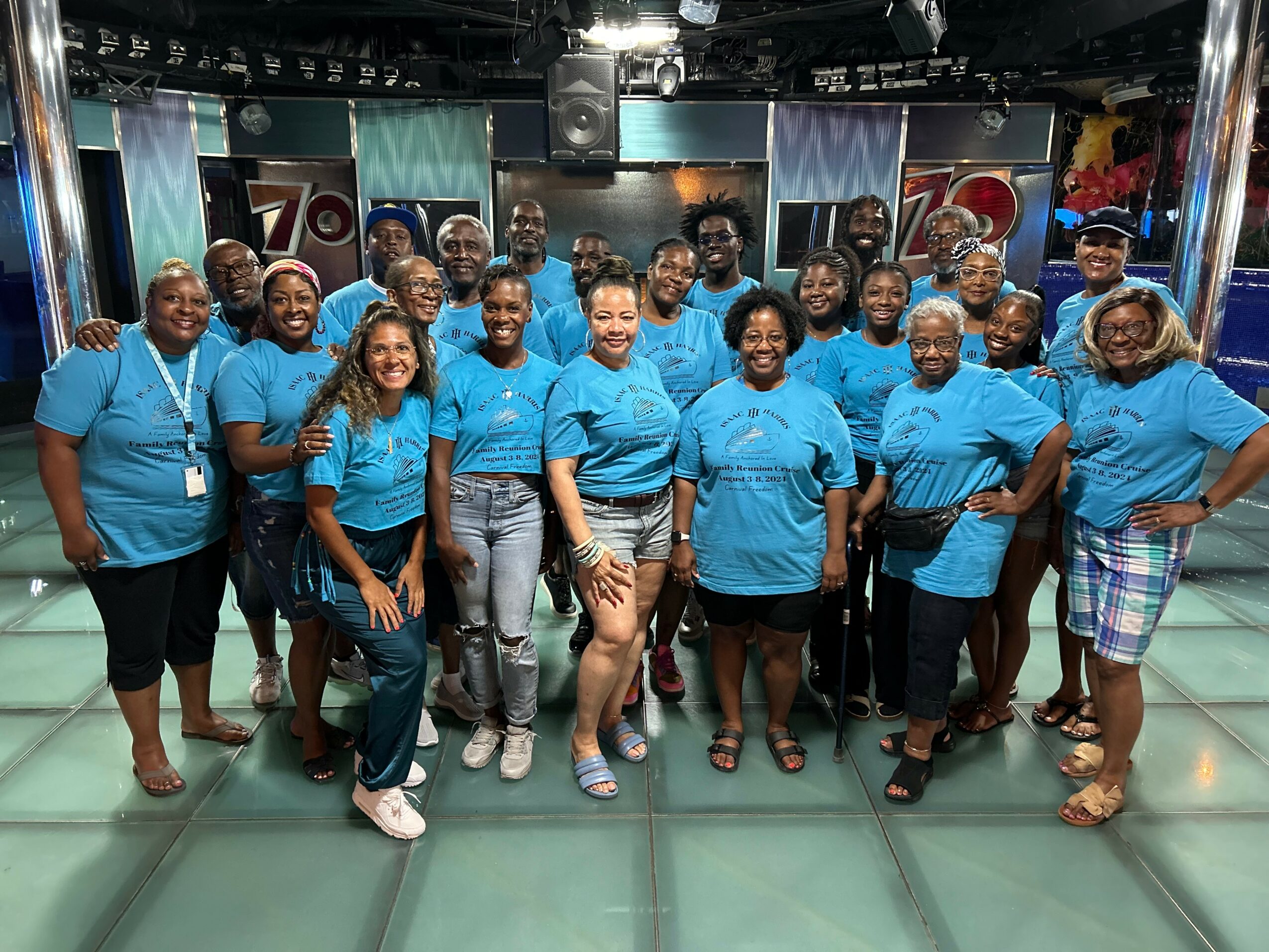 Group of people wearing matching blue shirts, smiling, standing indoors with stage and screens in background.