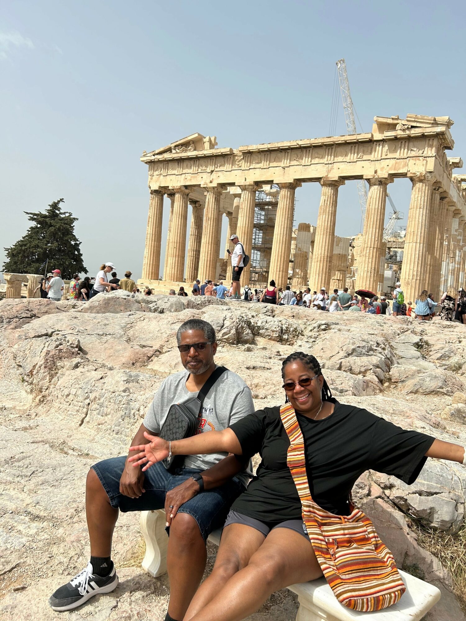 Two people sitting on rocks in front of an ancient Greek temple with columns, with many visitors in the background.