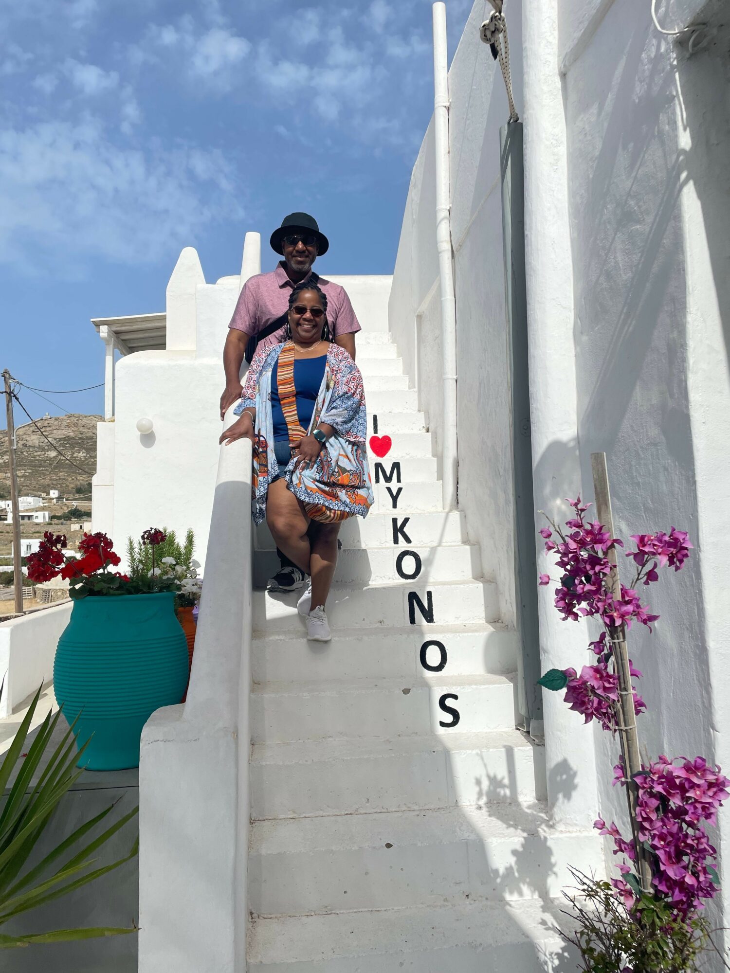 Person in colorful clothing walking on a sidewalk with white walls and pink flowers, sunny sky in background.