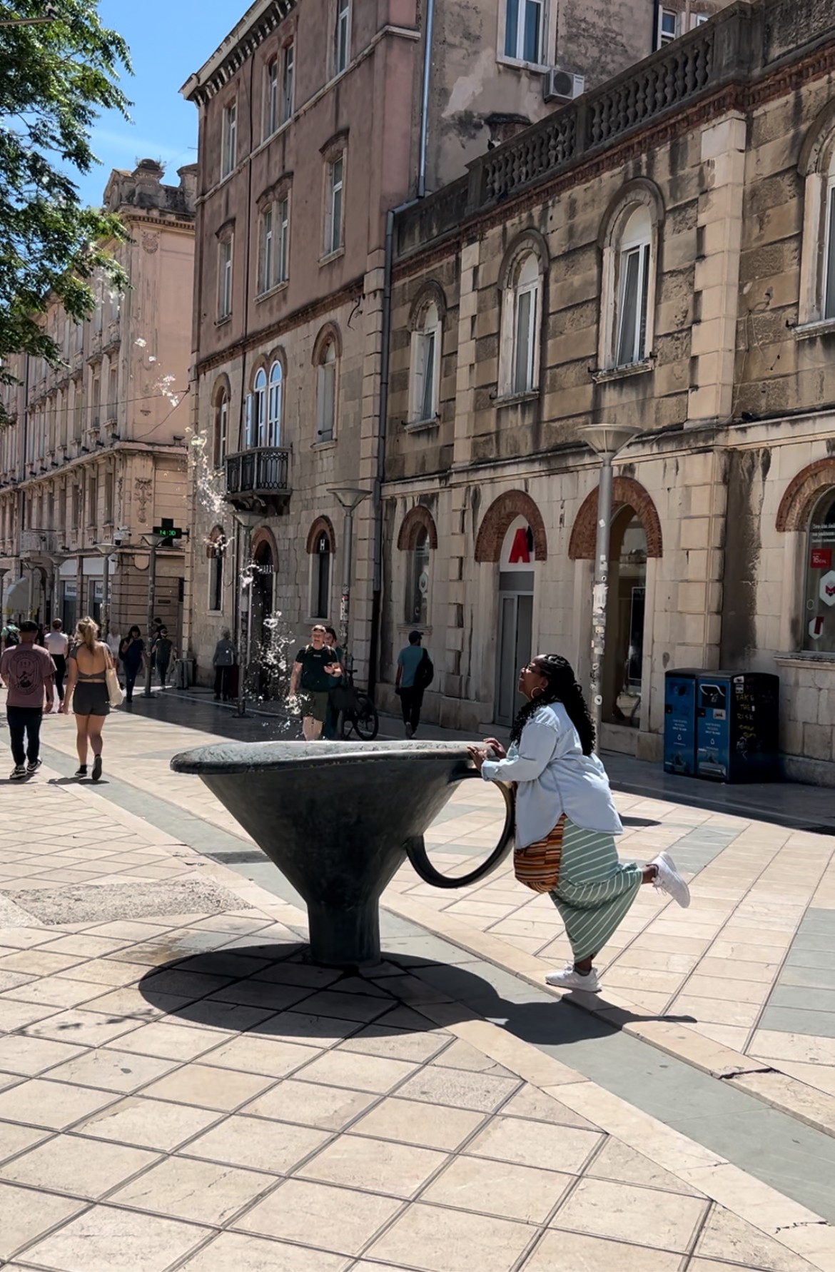 Person leaning on a large, black, bowl-shaped sculpture in a city square with historic buildings in the background.