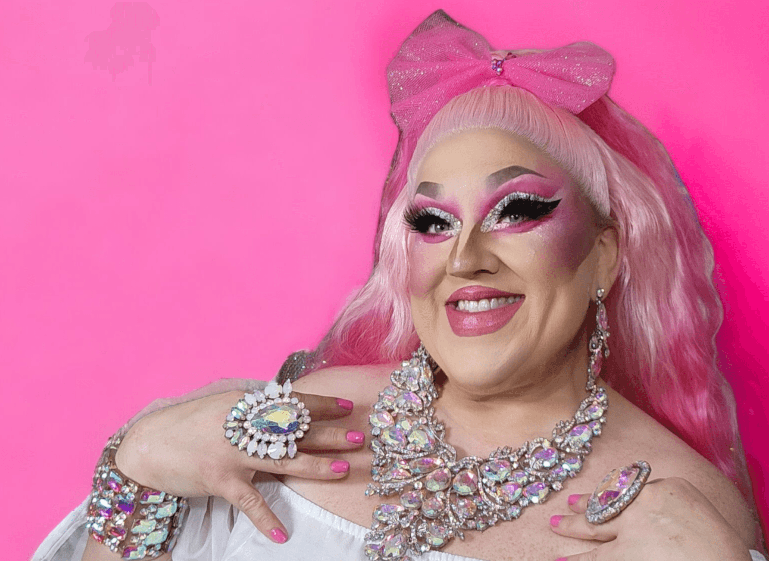 Person with pink hair, large jewelry, and makeup, smiling against a pink background.