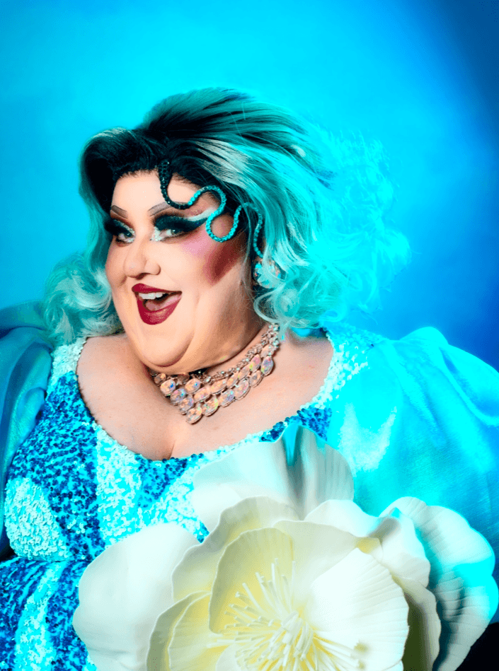 Person with vibrant teal and black hair, makeup, and jewelry, holding a large white flower, smiling brightly.