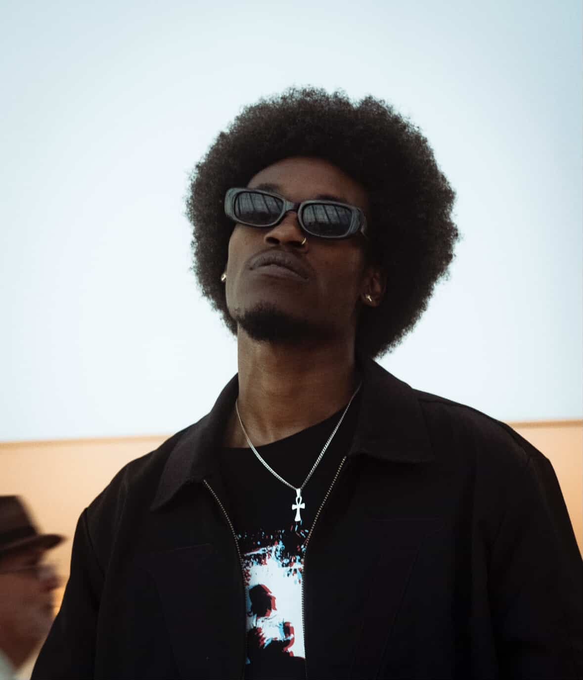Person with sunglasses and afro hairstyle wearing a black jacket and necklace, looking upward, against a light background.