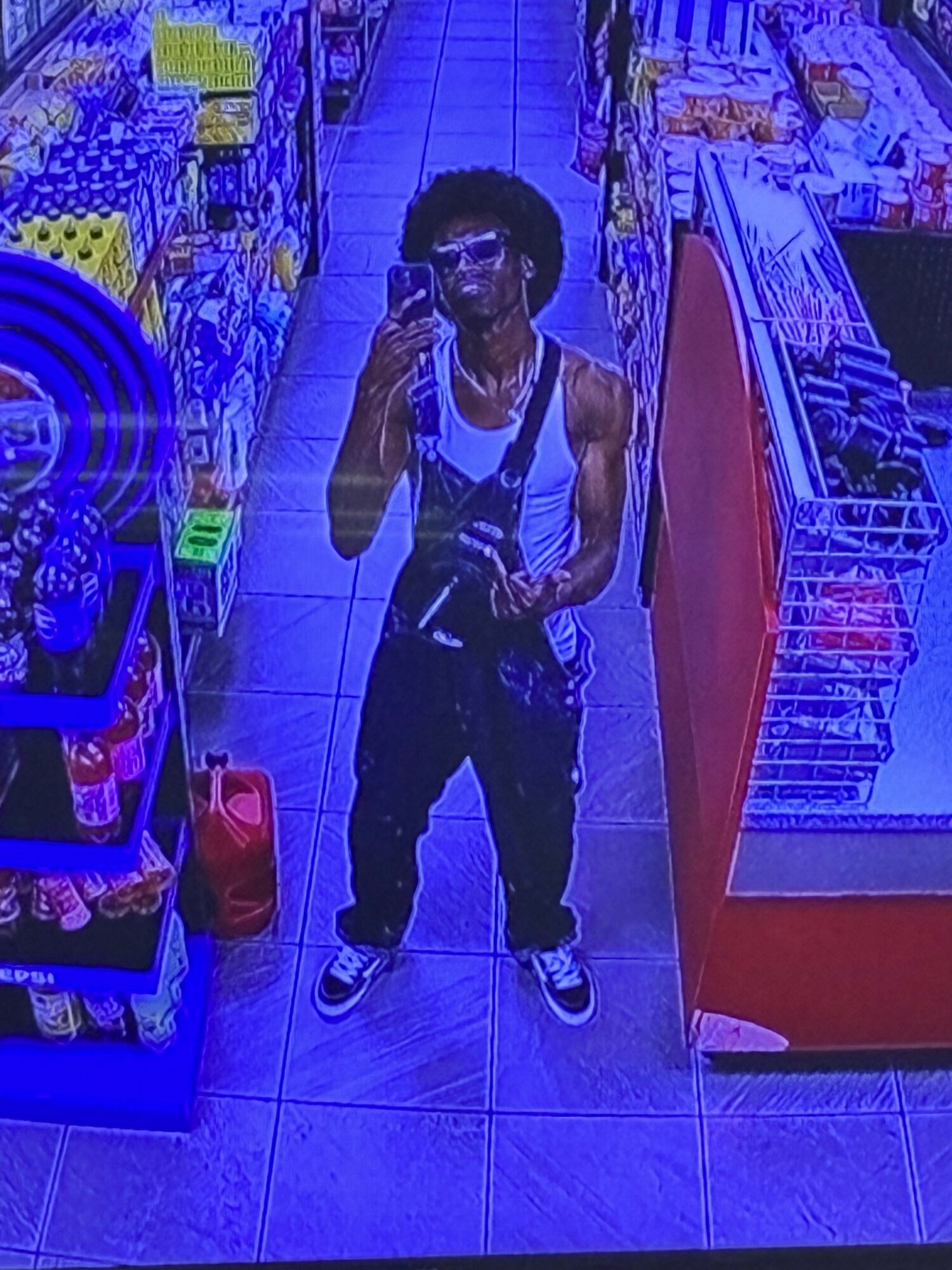 Child taking a selfie in a store aisle with shelves of products, holding a phone, wearing a white shirt and dark pants.