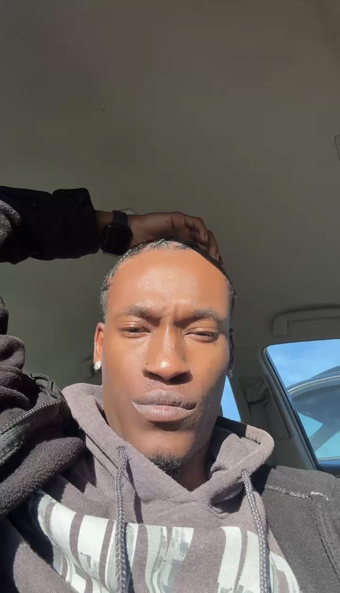 Young man with short hair and a hoodie, sitting in a car, looking at the camera, sunlight on face.