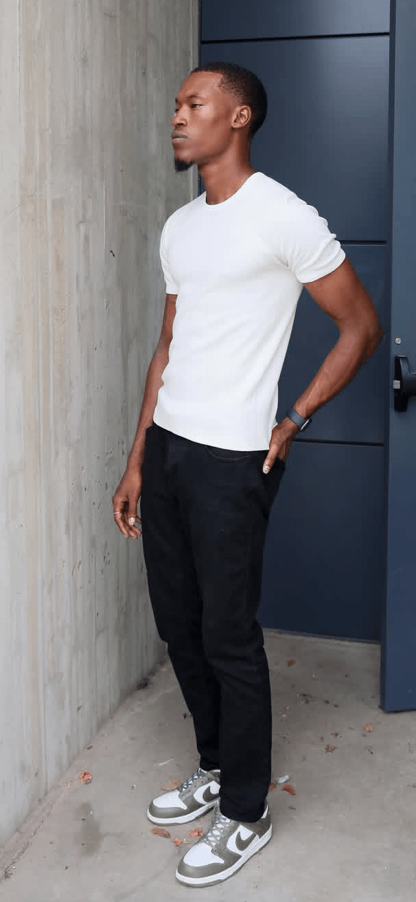 Young man standing sideways against a concrete wall, wearing a white t-shirt, black pants, and sneakers.