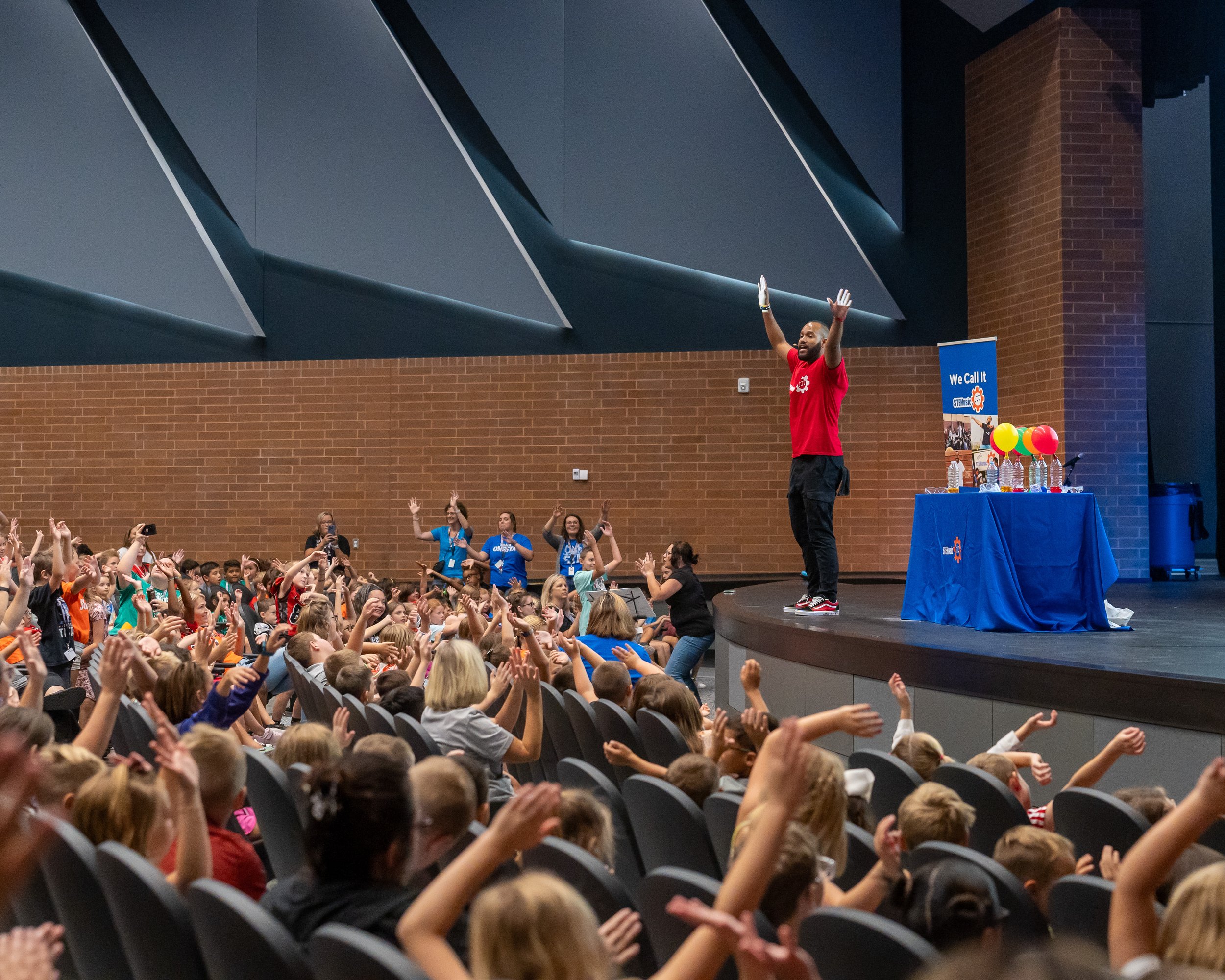 Person on stage with raised arm in front of large audience in auditorium, with a table and colorful balloons nearby.