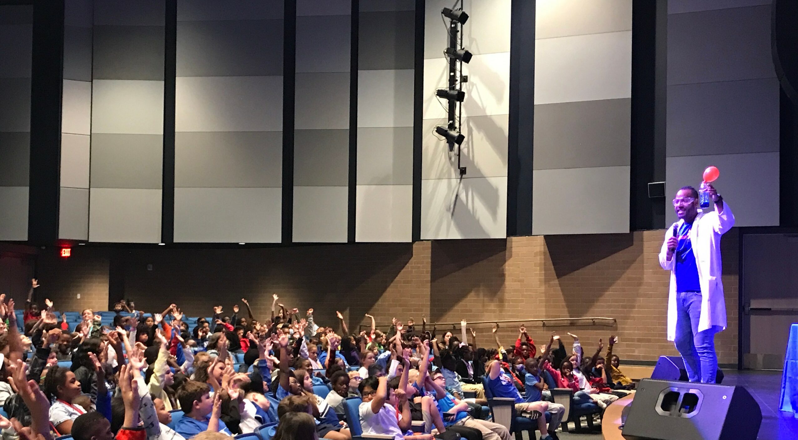 Speaker on stage with audience raising hands in a large auditorium, high ceiling with panels, speaker holding microphone.