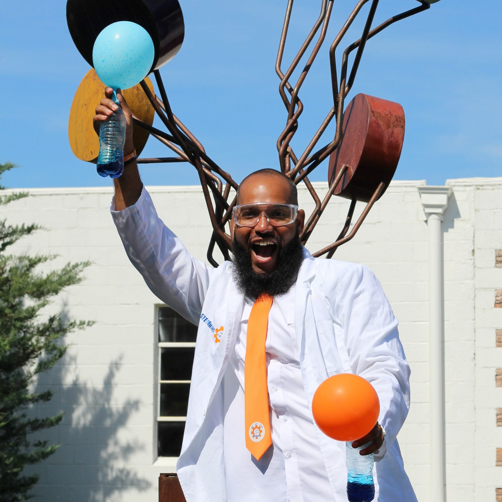 Man with glasses and beard smiling, holding a blue balloon and an orange ball, outdoors with a white building and blue sky