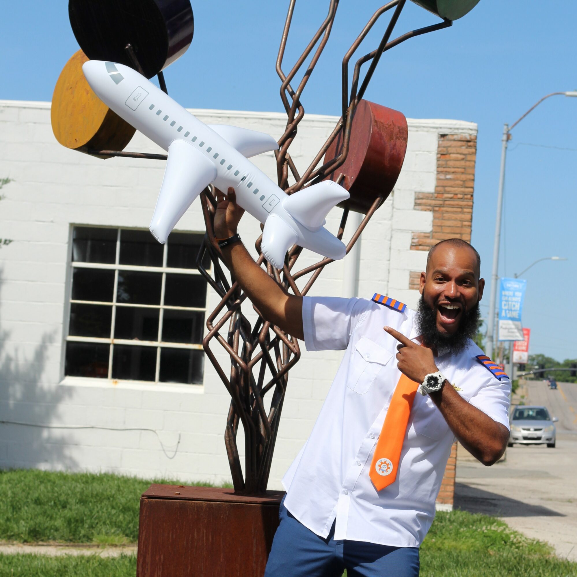 Man in uniform pointing at a sculpture of an airplane and trees outdoors, smiling happily.