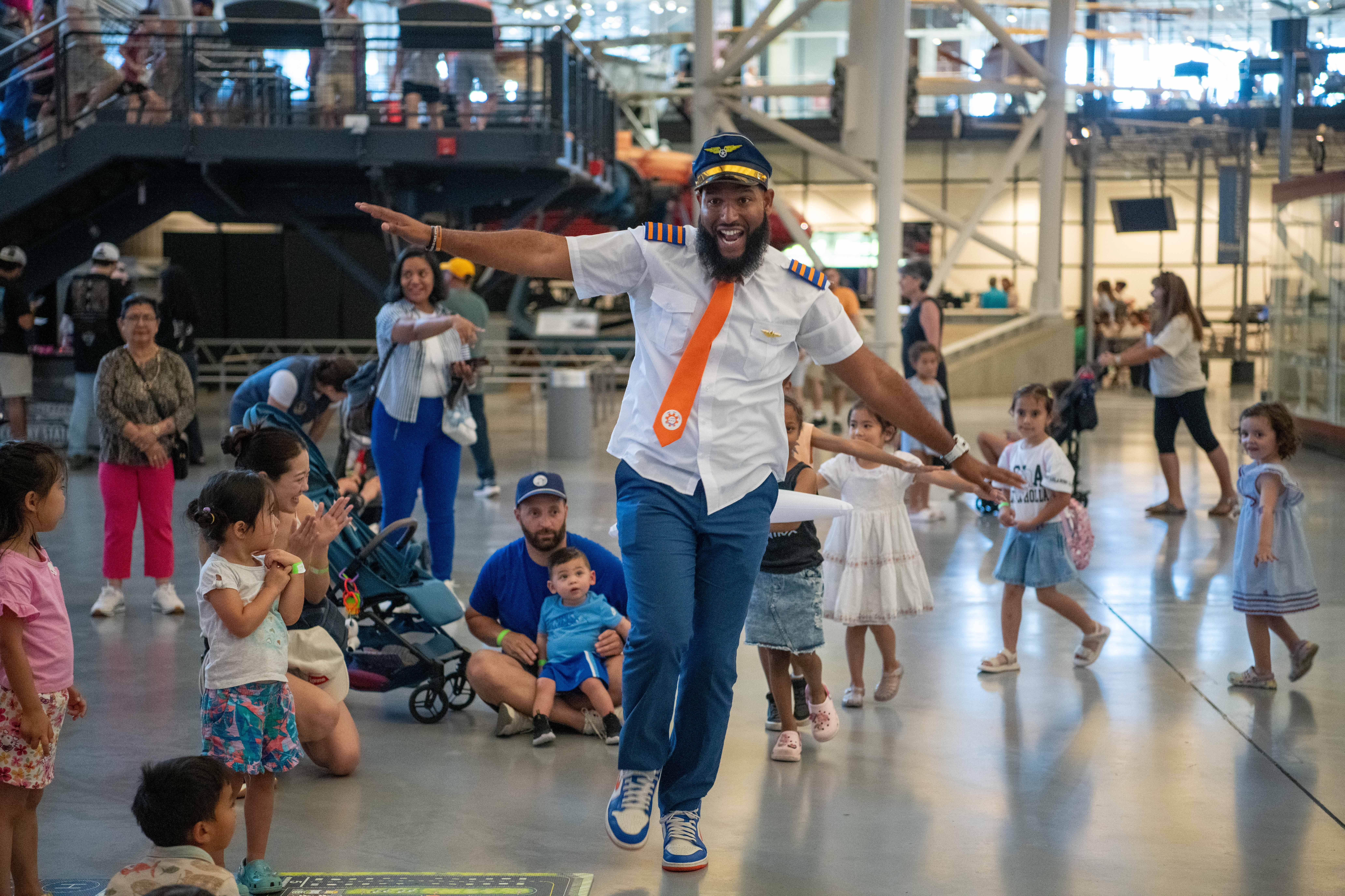 Man with a beard and glasses dancing with children in an indoor space, some children clapping and playing, people in background.