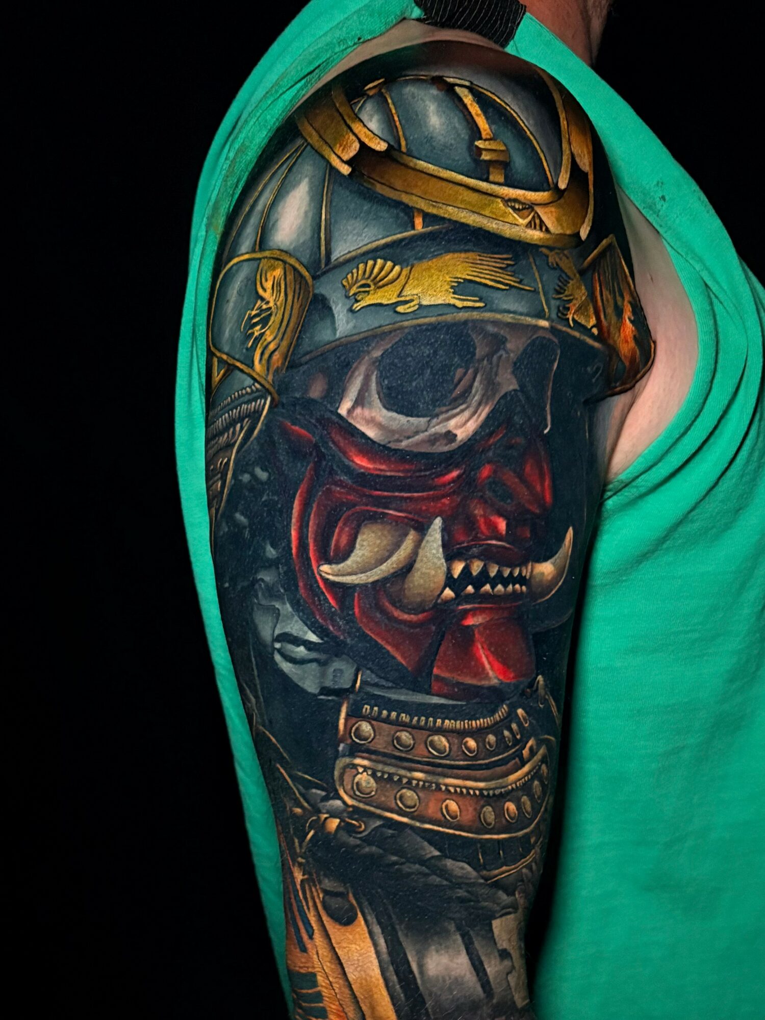 Tattoo of a samurai helmet, dragon, and fierce red face mask on person's upper arm, with green clothing partially visible.