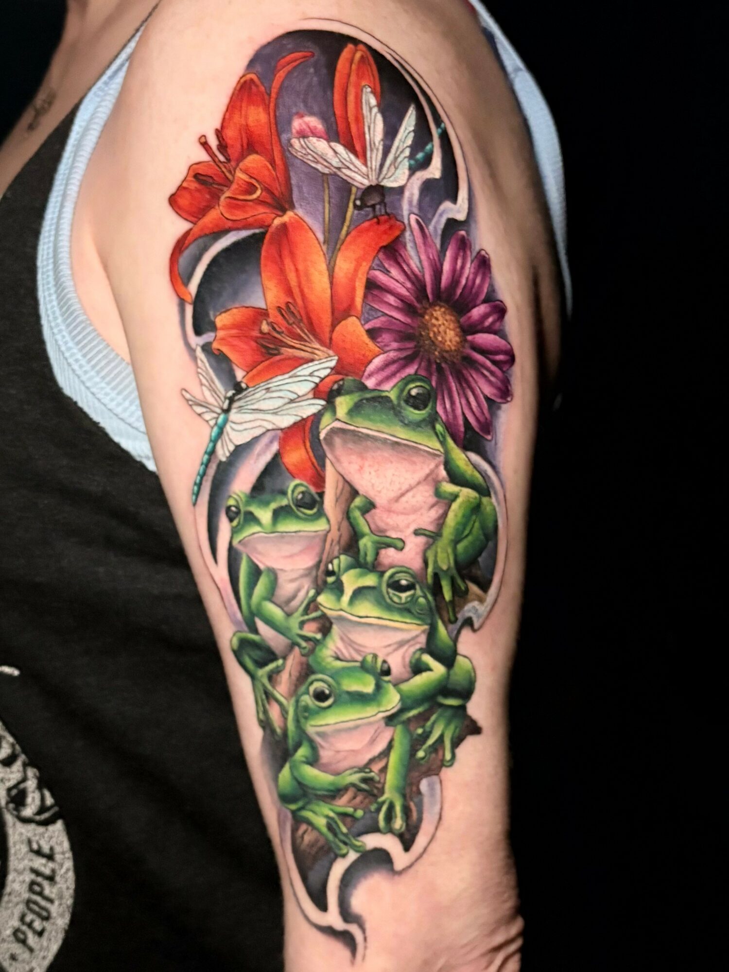 Colorful tattoo of lilies, a daisy, a frog, and dragonflies on a person's upper arm.