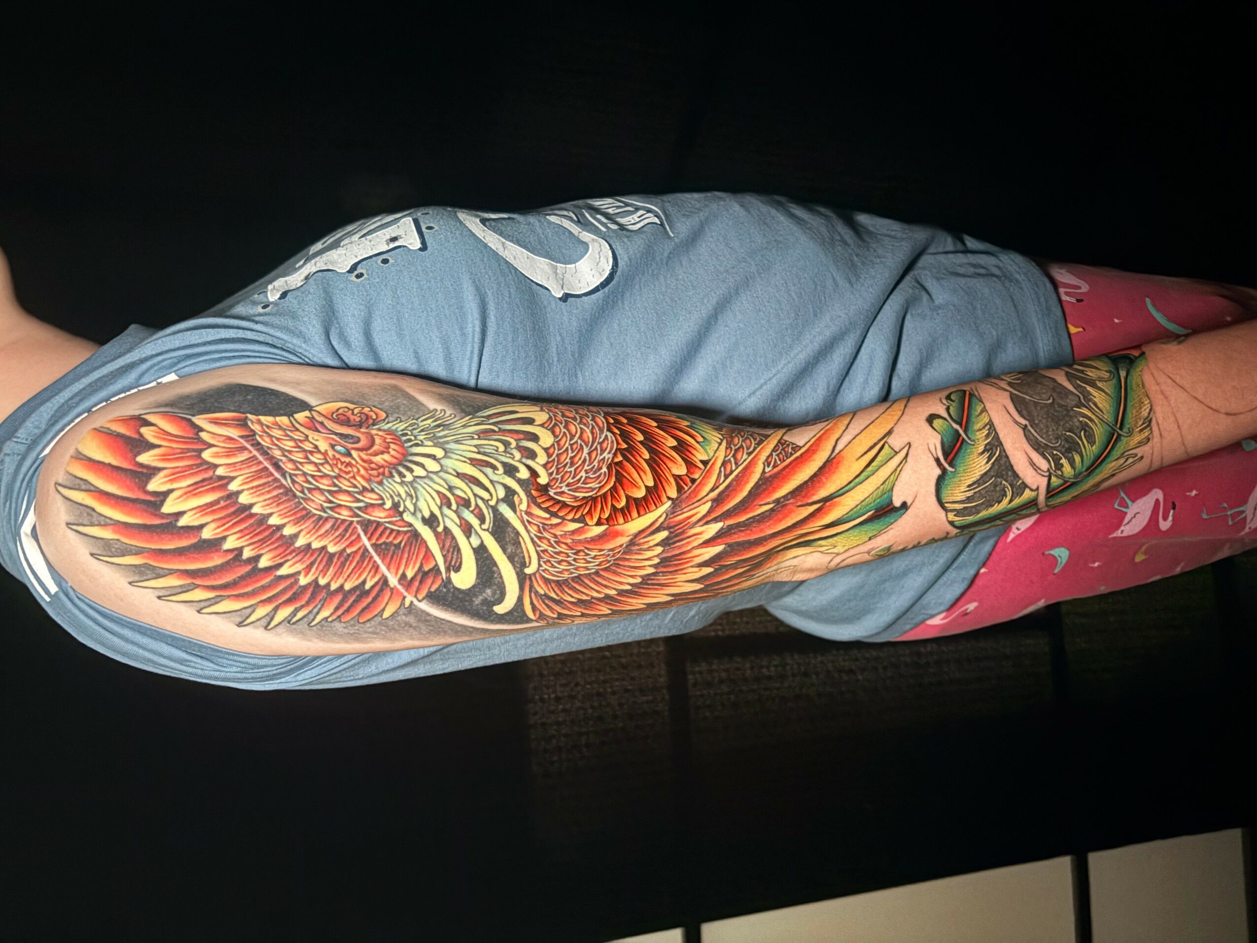 Person lying on side with colorful phoenix tattoo on arm, wearing a blue shirt, dark background.