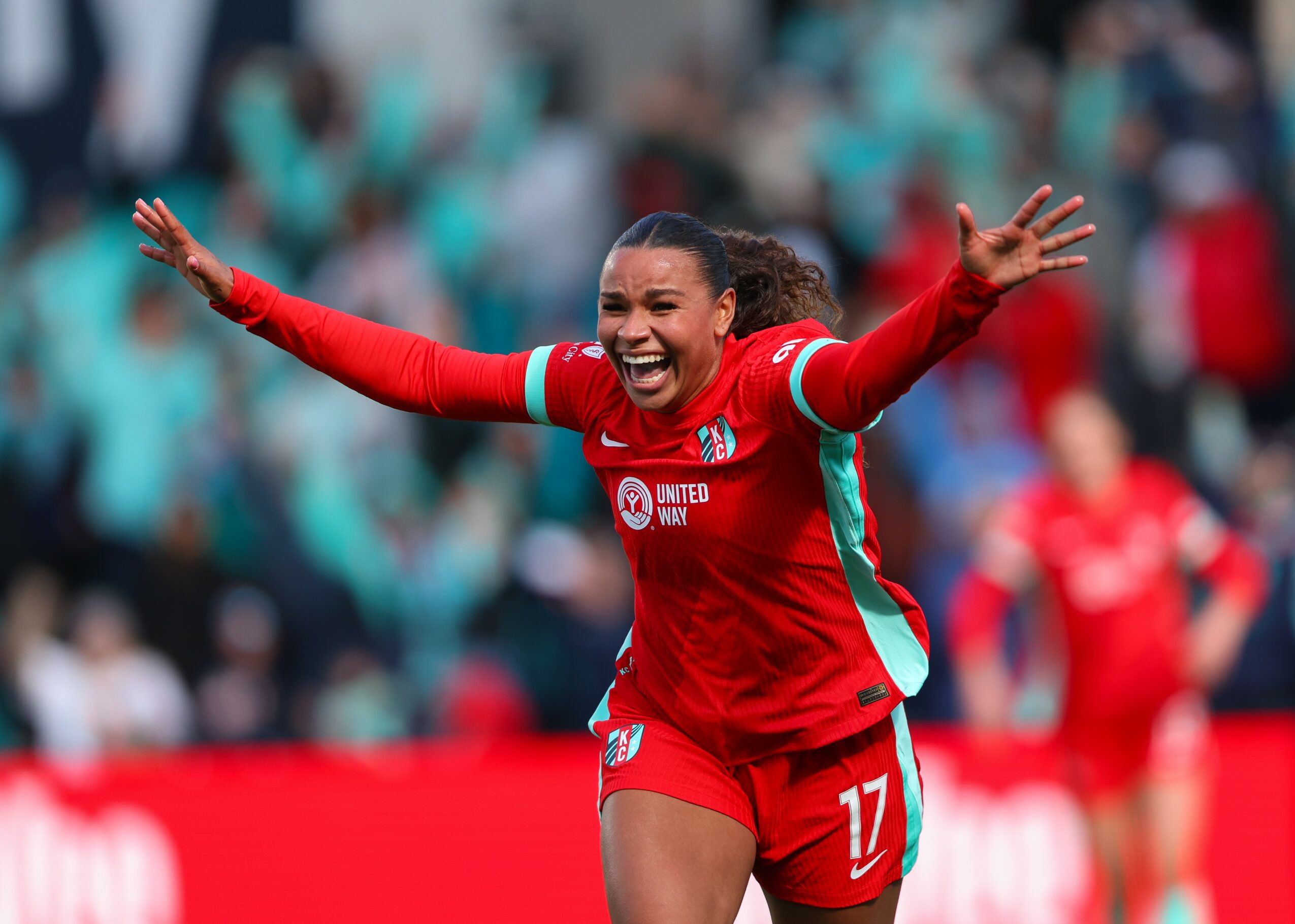 Female soccer player celebrating with arms outstretched, smiling, wearing red uniform with number 17, in a stadium.