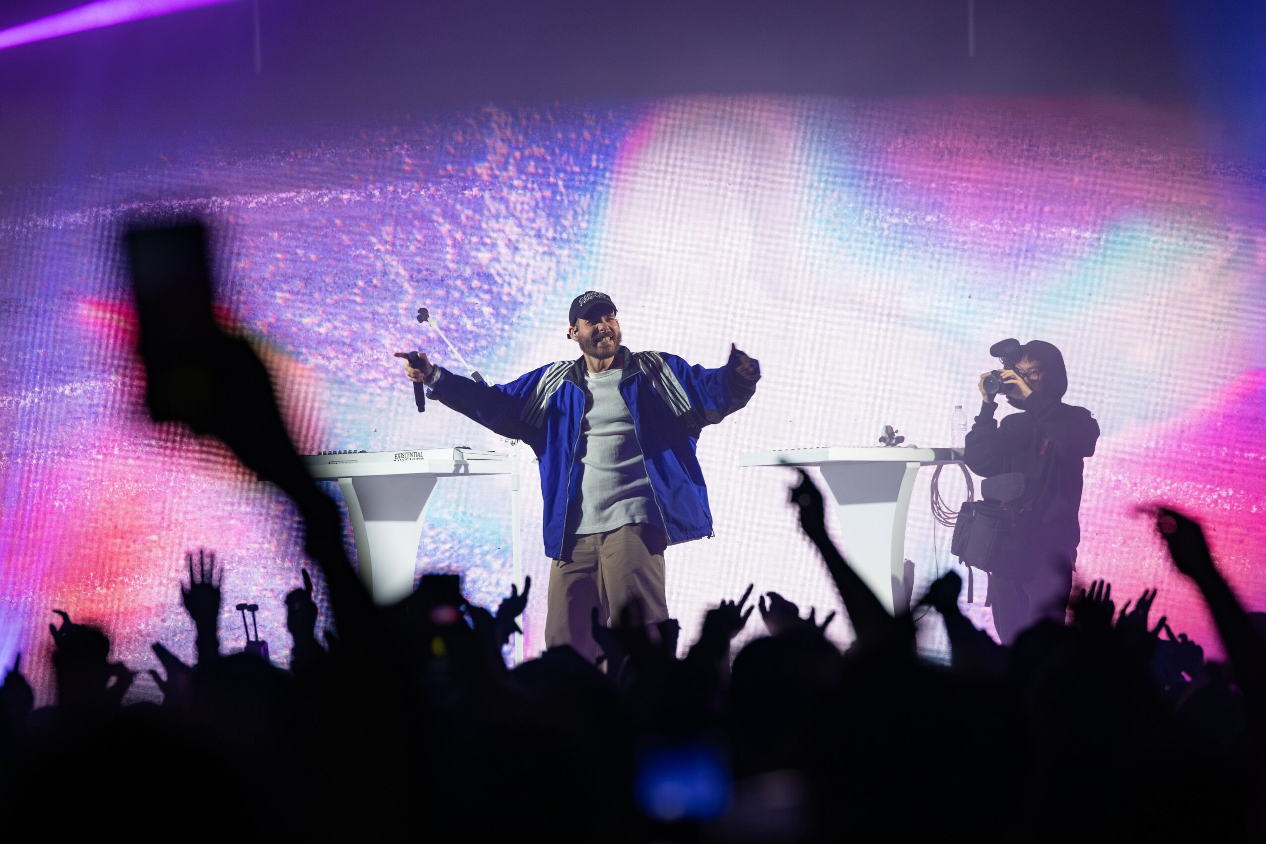 Performer on stage with arms outstretched, colorful background, audience with raised hands, and a person with camera on right.