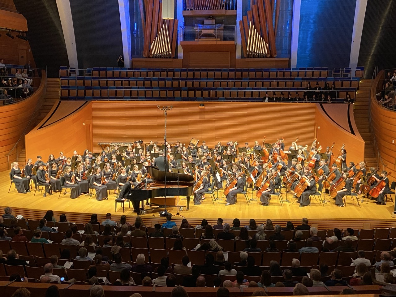 Orchestra performing on stage with a conductor, piano, and audience in a concert hall.