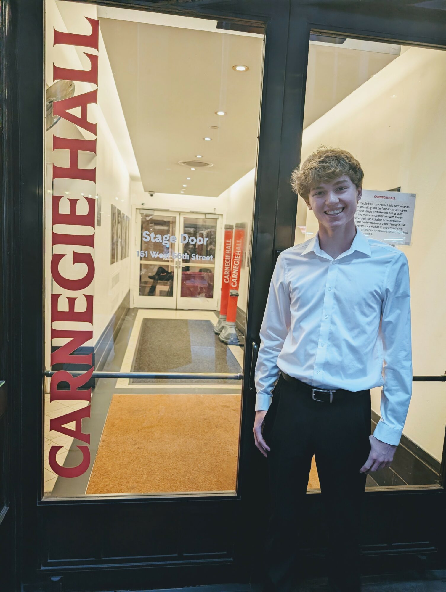 Young man in white shirt and black pants standing inside Carnegie Hall lobby, smiling, with Carnegie Hall sign visible outside.