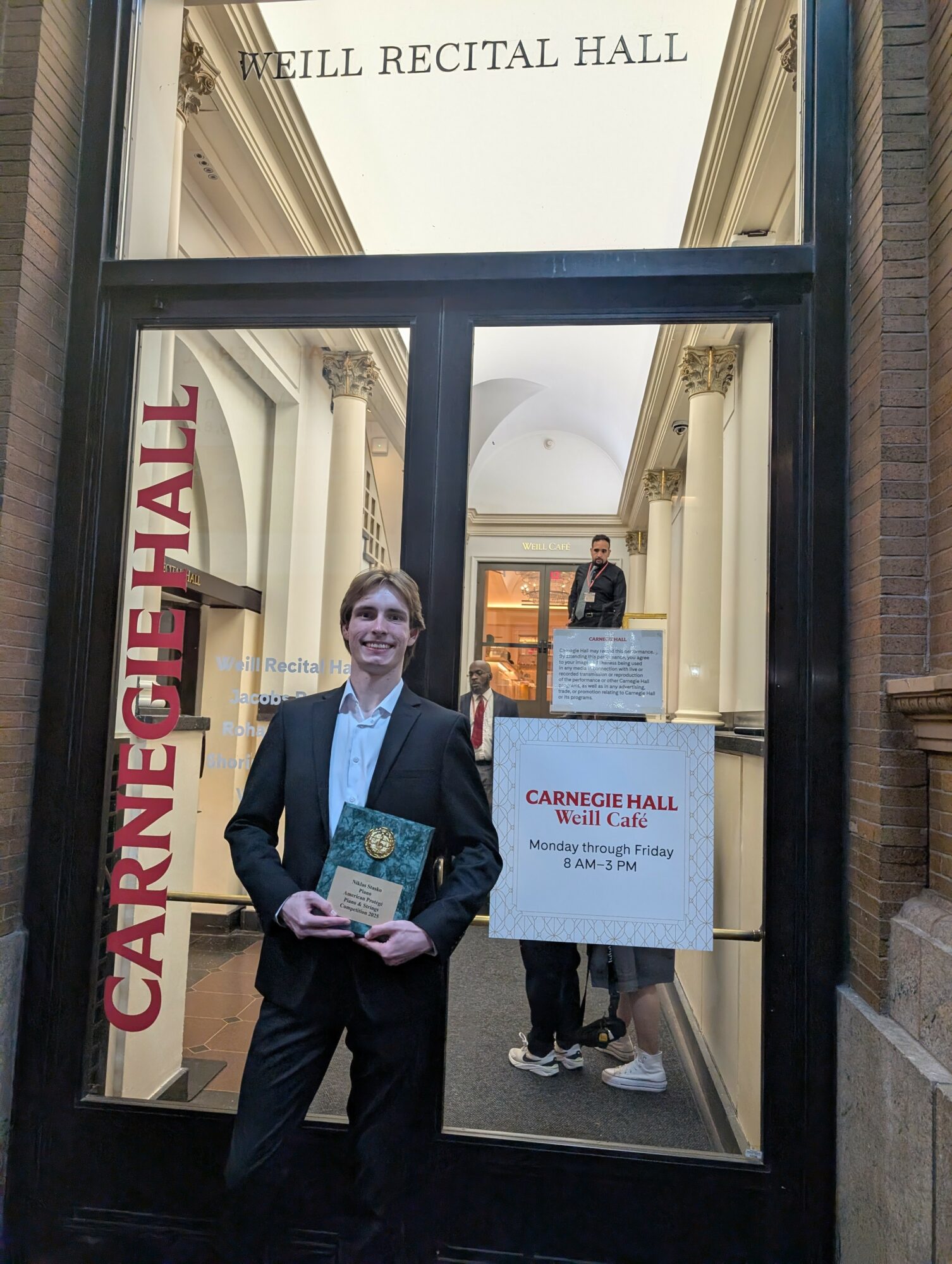 Person in formal attire holding a book and standing in front of Carnegie Hall entrance, with interior visible through glass door.