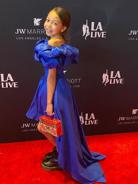 Young girl in a blue dress holding a red handbag, standing on a red carpet with a black backdrop.