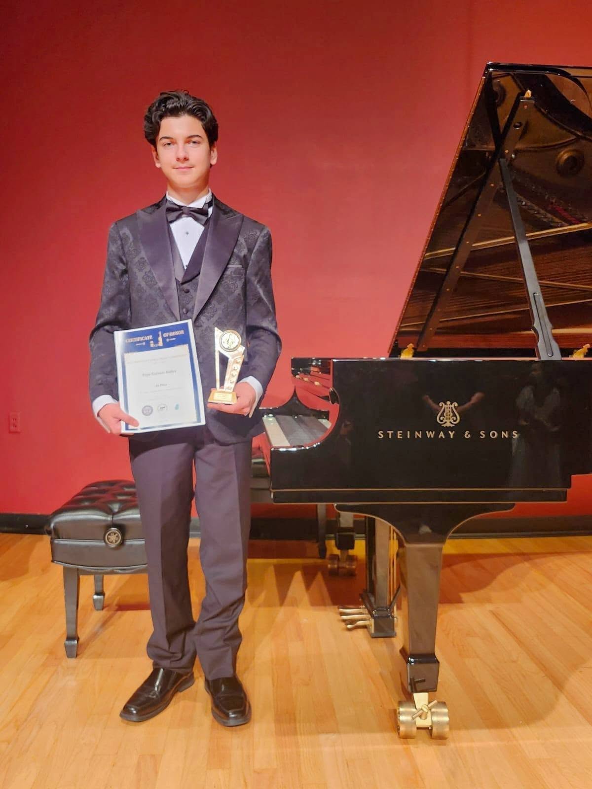 Young person in formal suit holding a certificate and trophy, standing next to a grand piano on stage.