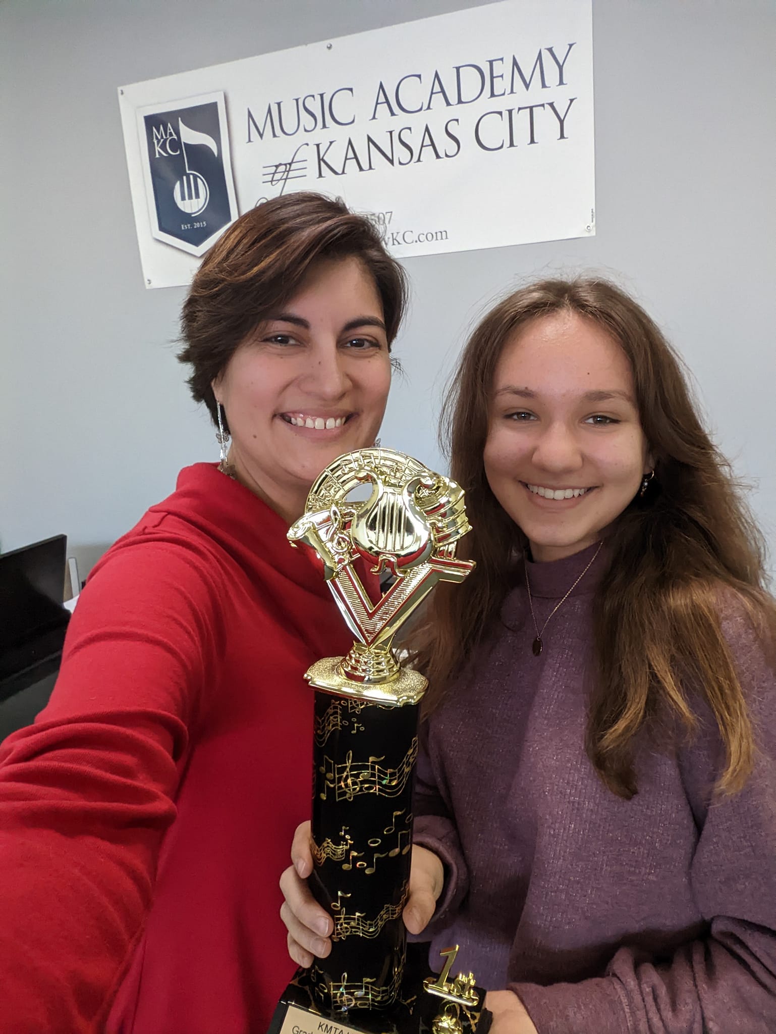 Two smiling women holding a trophy in front of a sign that reads 'Music Academy of Kansas City.'