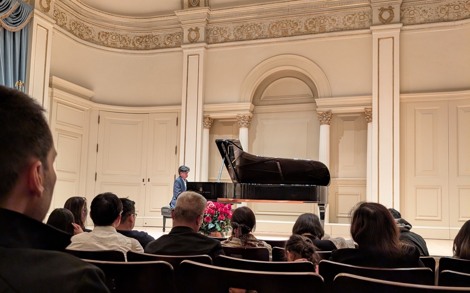People seated in an auditorium watching a person perform on a grand piano on stage, with ornate architecture in the background.