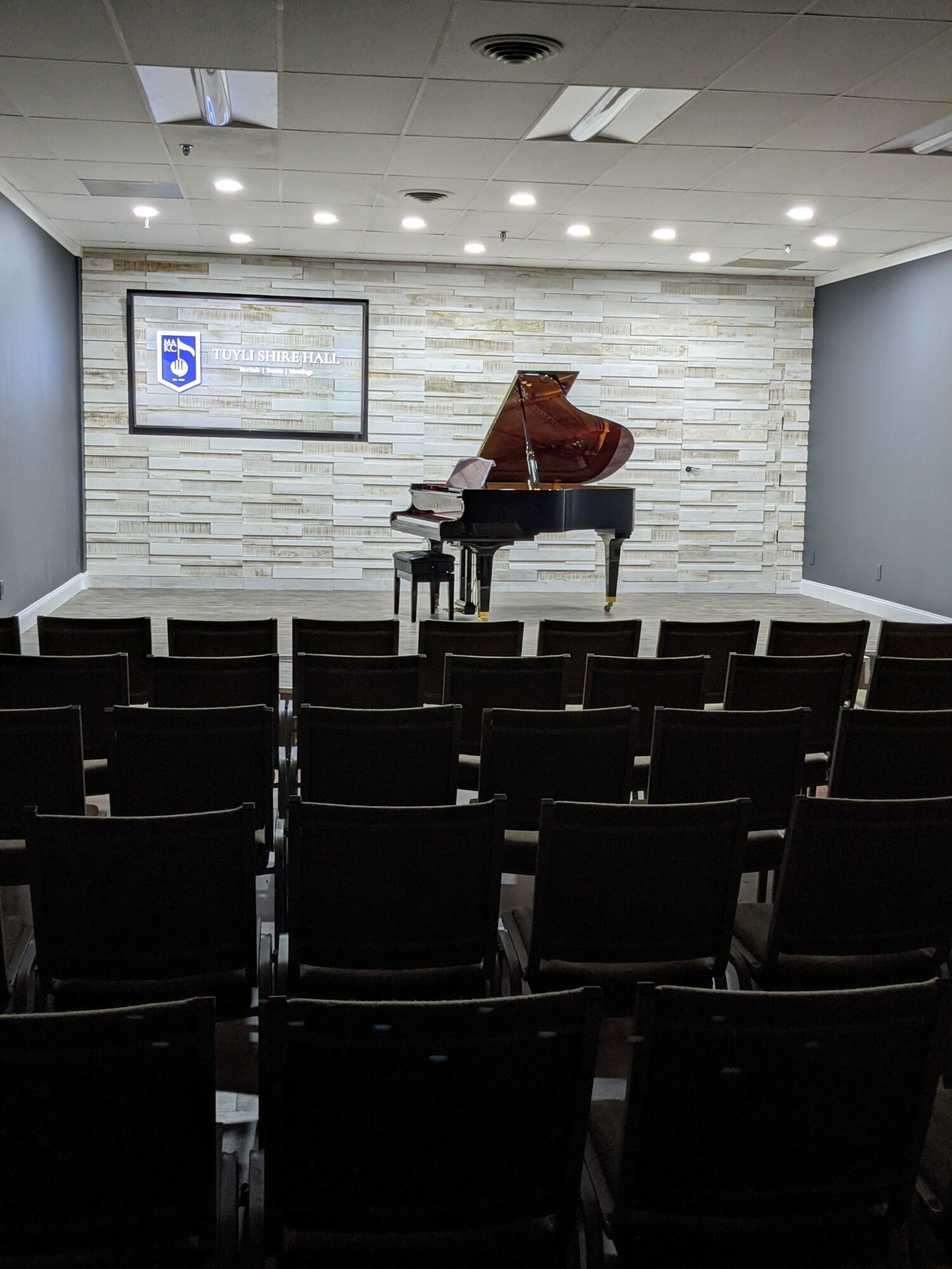 Empty auditorium with a grand piano on stage, rows of chairs, and a screen on the wall, neutral lighting.