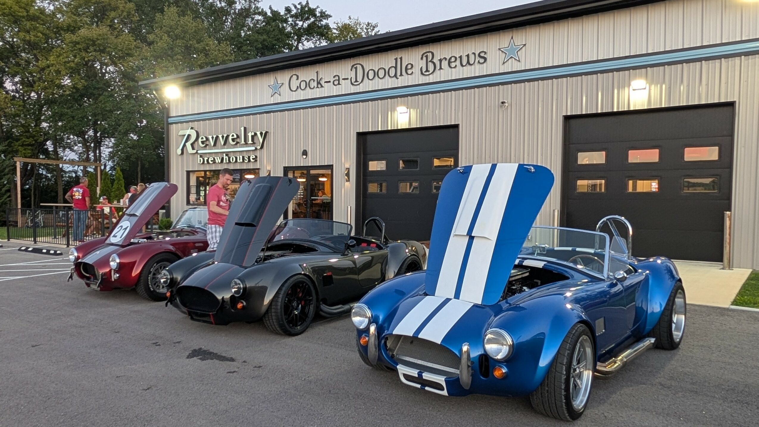 Four vintage cars parked in front of a building with a sign reading 'Cock-a-Doodle Brews' and 'Jewelry'.