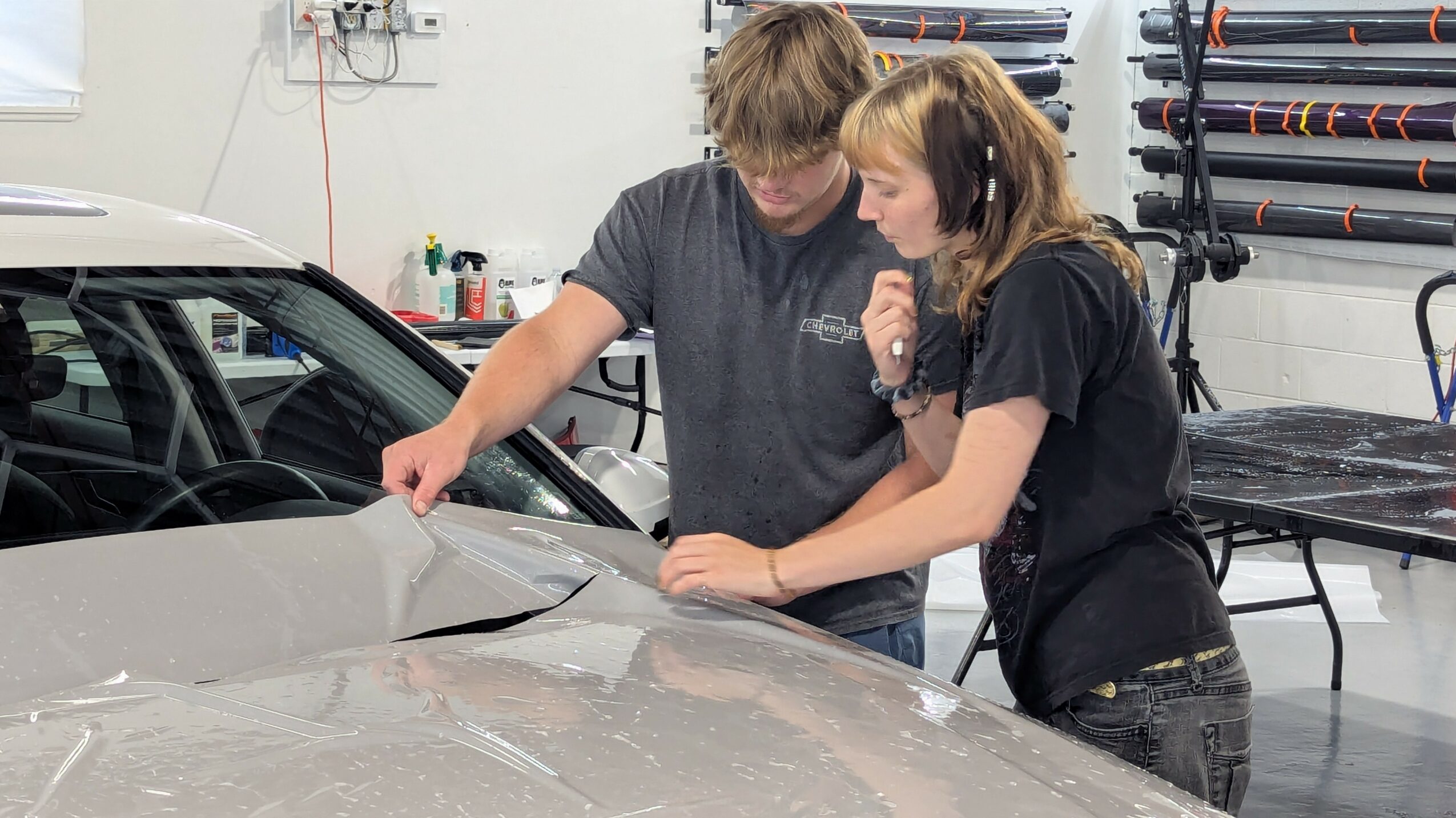 Two people examine a damaged car in a workshop, discussing the impact area on the vehicle's front. Workshop tools are visible in the background.