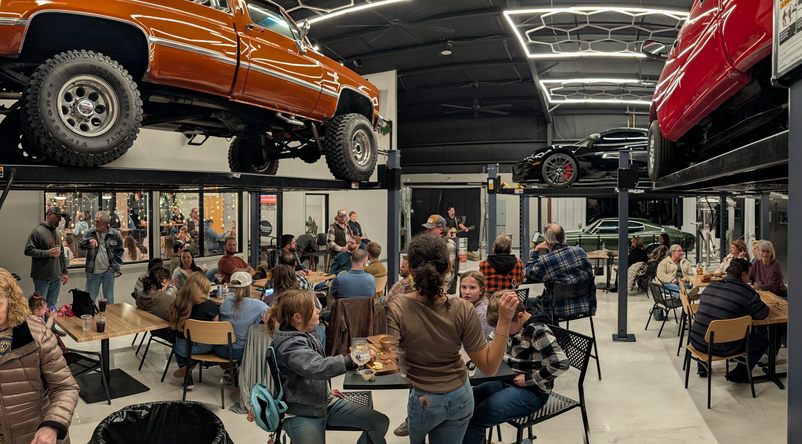 People gather and sit at tables in a large indoor space with cars displayed on elevated platforms above them.
